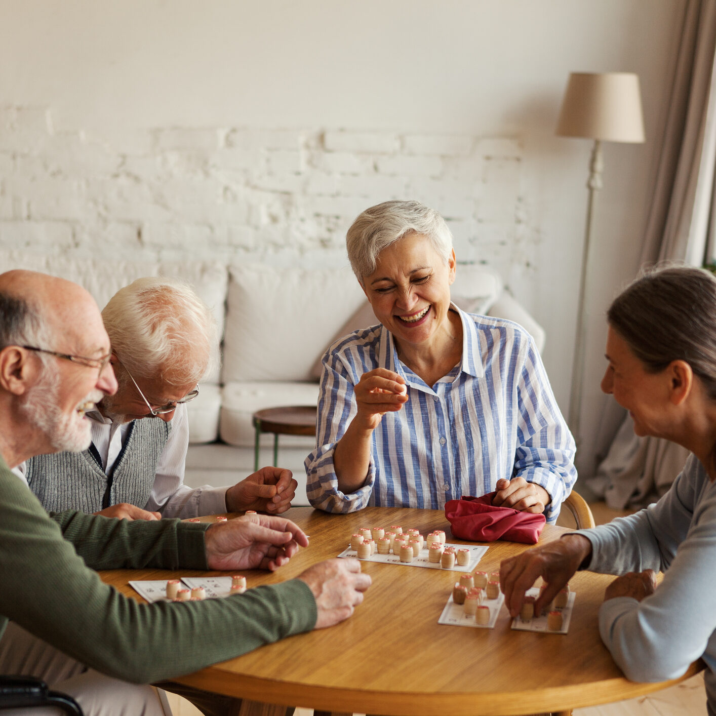 Group of four cheerful senior people, two men and two women, having fun sitting at table and playing bingo game in nursing home