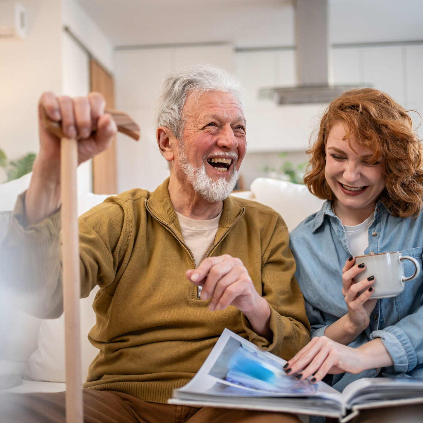 Senior man using a walking cane sits on the sofa with a young woman, both laughing and enjoying tea while looking through a photo album, sharing cherished memories and moments of joy